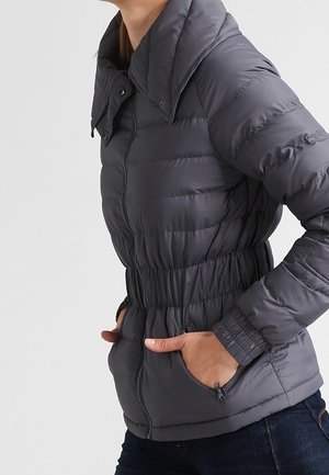 Personne portant une doudoune gris foncé avec taille élastique et mains dans les poches latérales zippées, montrée du cou aux hanches.