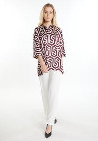 Blusa geometrica bordeaux e bianca con colletto e maniche a 3/4, abbinata a pantaloni bianchi e décolleté nere a punta.