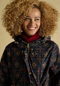 Ūdensizturīga melna jaka ar ģeometrisku zīmējumu zeltā un zilā krāsā. Iekļauj rāvējslēdzēju, kapuci un sarkanu ribotu apkakli.