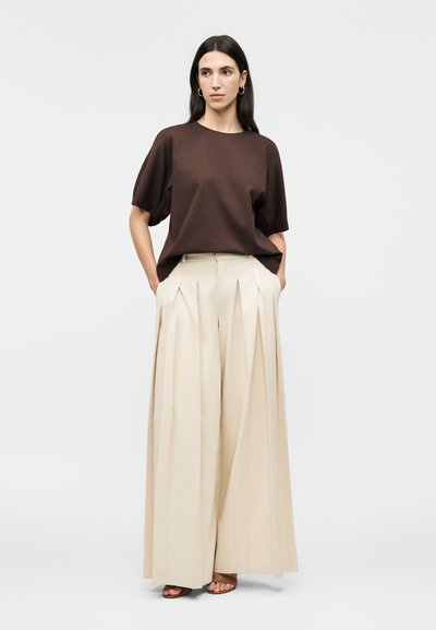 Γυναίκα με σκούρο καφέ μπλούζα με κοντά μανίκια, εκρού πλισέ παντελόνι ψηλόμεσο με φαρδιές παντελόνες και καφέ σανδάλια ανοιχτά στο πόδι, στέκεται με τα χέρια στις τσέπες.
