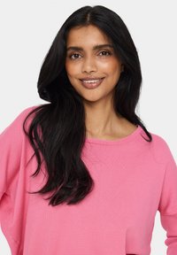 Rozā adīta džemperis ar brīvu piegriezumu, plašu apkakli un garām piedurknēm. Mīksta tekstūra ar smalku ribošanu uz piedurknēm un apakšmalas.