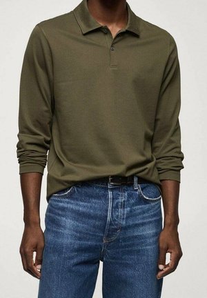 Mies, joka pukeutunut oliivinvihreään pitkähihainen poolopaitaan, joka on työnnetty sinisiin farkkuihin mustan vyön kanssa, seisoo yksinkertaista vaaleaa taustaa vasten.