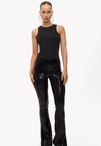 Femme portant un haut noir sans manches et un pantalon taille haute en sequins noirs brillants, debout devant un fond blanc uni, avec de longues boucles d'oreilles.