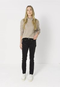 Femme debout portant un pull à col roulé beige, un pantalon noir et des baskets blanches sur un fond blanc uni.