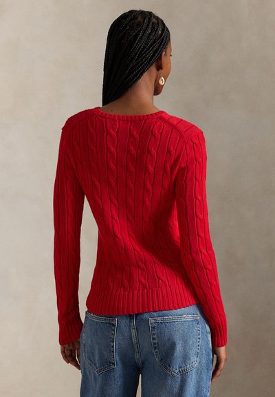 Κόκκινο πουλόβερ με πλέξη cable-knit, μακριά μανίκια, ριμπ τελείωμα και κλασικό στρογγυλό λαιμό. Φορεμένο πάνω από μπλε τζιν παντελόνι, παρουσιάζοντας λεπτομέρειες υφής υφάσματος.