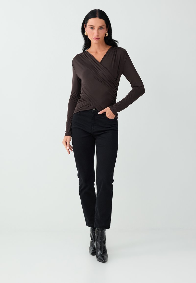 Vrouw met lang donker haar die een donkerbruine wikkelstijl top met lange mouwen, zwarte broek en zwarte enkellaarzen draagt, staand tegen een effen achtergrond.