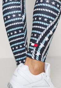 Leggings con una base blu navy e un motivo di corde blu e stelle bianche. Presentano una piccola bandiera rossa, bianca e blu alla caviglia. Indossati con sneakers bianche.