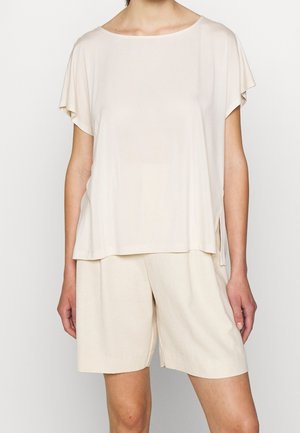 Maglietta corta a maniche con un color beige chiaro, dal fit rilassato, in tessuto morbido, con scollatura rotonda e spacchetti laterali, abbinata a pantaloni corti larghi dello stesso colore.
