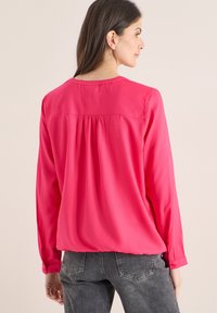 Blouse à manches longues en tissu rose vif à texture lisse, avec un pli sur le yoke arrière et des poignets boutonnés.
