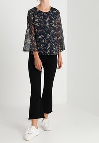 Blusa trasparente blu navy con motivo floreale e maniche a campana abbinata a pantaloni neri a zampa e sneakers bianchi. Tessuto dalla texture liscia.