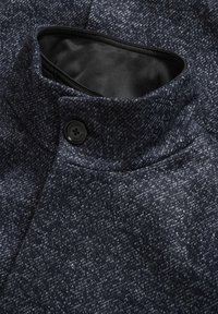 Tessuto a fantasia blu scuro e grigio con un alto colletto, dotato di un bottone nero e una fodera interna liscia. Dettaglio ravvicinato del cappotto.