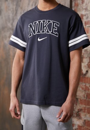 Homme portant un t-shirt Nike bleu marine avec des rayures blanches sur les manches et un pantalon de survêtement gris clair devant un mur en briques.