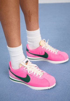 Ben iført hvide ribstrikkede sokker og knaldrosa Nike-sneakers med mørkegrøn swoosh på blåt tæppe.