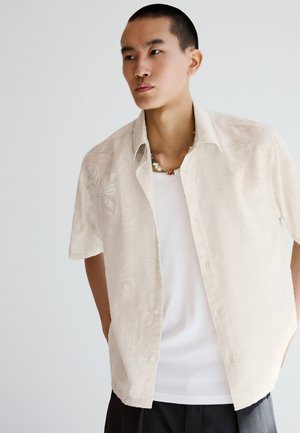 Junger Mann trägt ein offenes beige gemustertes kurzärmeliges Hemd, weißes Tanktop, schwarze Hose und bunte Perlenkette, schaut zur Seite.