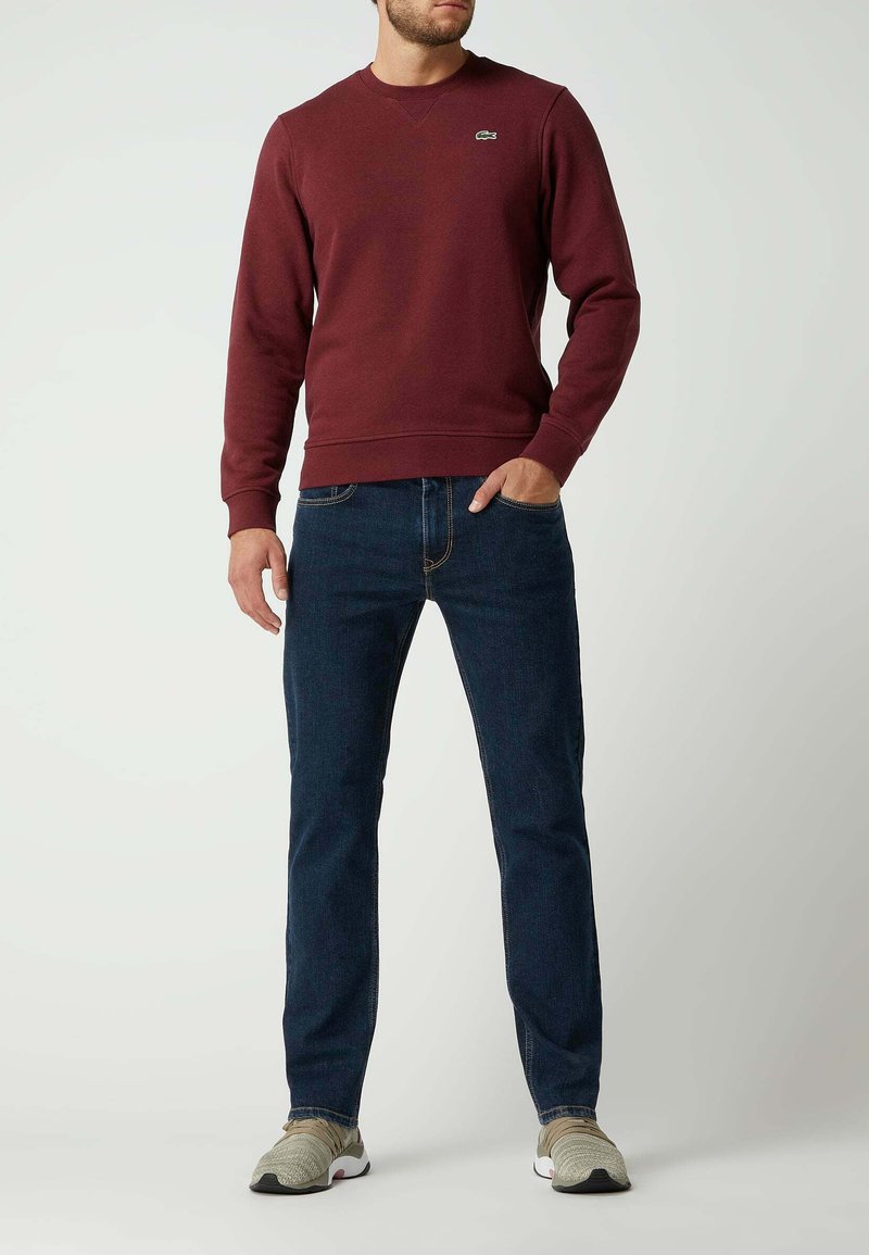 Burgundy sweatshirt with crew neck and ribbed cuffs, paired with dark blue jeans. Model wears light gray sneakers with textured pattern.