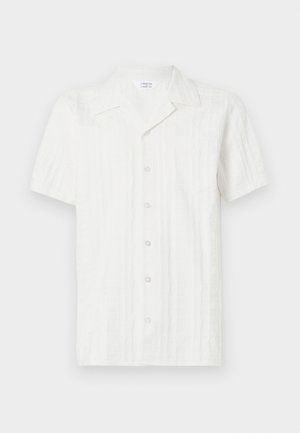 Valkoinen lyhythihainen paita, jossa on teksturoitu kuvio, napitus edessä ja yksi tasku vasemmalla puolella. Valmistettu kevyestä kankaasta.