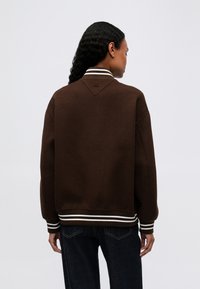 Persona con cabello largo y rizado, vistiendo una chaqueta marrón con puños y dobladillo de rayas blancas, de espaldas frente a un fondo sencillo.