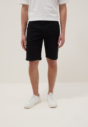 Homme portant un short noir jusqu'aux genoux, un t-shirt blanc et des baskets blanches, debout sur un fond clair uni.