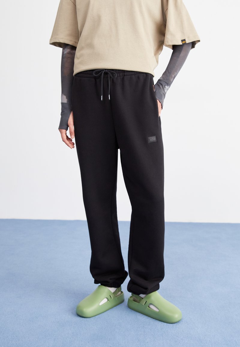 Persoon in een beige T-shirt, zwarte joggingbroek met een trekkoord, lange grijze mouwen eronder en lichtgroene instappers, staand op een blauwe vloer.