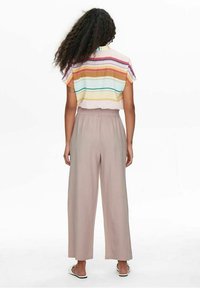 Kvinna med långt lockigt hår som bär en kortärmad, flerfärgad randig skjorta, beige vida byxor och vita sandlekskor, stående med ryggen mot kameran.