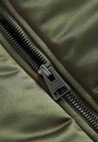 Tessuto verde oliva con texture liscia, dotato di una cerniera argentata e accessori in metallo. La cerniera è parzialmente aperta, mostrando l'imbottitura.