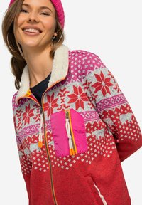 Giacca in fleece rossa e rosa con motivi floreali, accentata di bianco, colletto in pelliccia sintetica e tasca con cerniera. Presenta un tirazip arancione.
