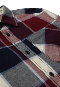 Zoom su una camicia a quadri con quadrati rossi, blu navy, grigi e crema, dotata di bottoni neri e una tasca sul petto.