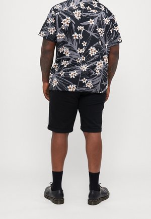 Mann trägt schwarzes, geblümtes Kurzarmhemd, schwarze hochgekrempelte Shorts, schwarze Socken und schwarze Schnürschuhe, steht mit dem Rücken zur Kamera.