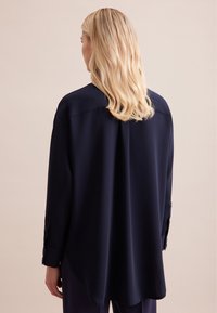 Camicia a maniche lunghe blu navy con vestibilità rilassata, tessuto morbido, polsini con bottone e orlo arrotondato, mostrata da dietro.