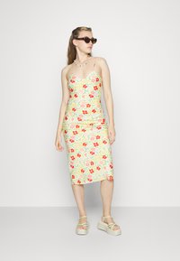 Vestido midi floral com alças finas. Fundo amarelo claro, padrão de flores vermelhas e verdes. Usado com sandálias verdes robustas.