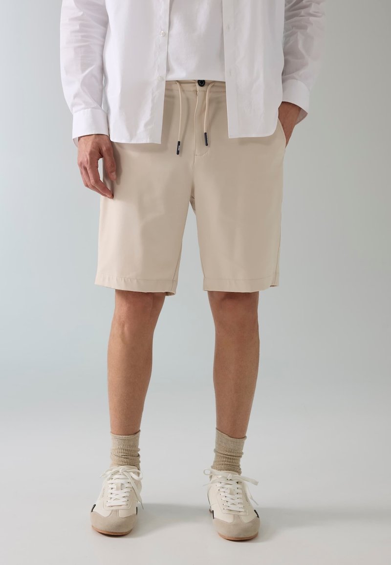 Homme portant un short beige à cordon, une chemise blanche boutonnée, des chaussettes beiges et des baskets blanches, debout devant un fond gris clair.