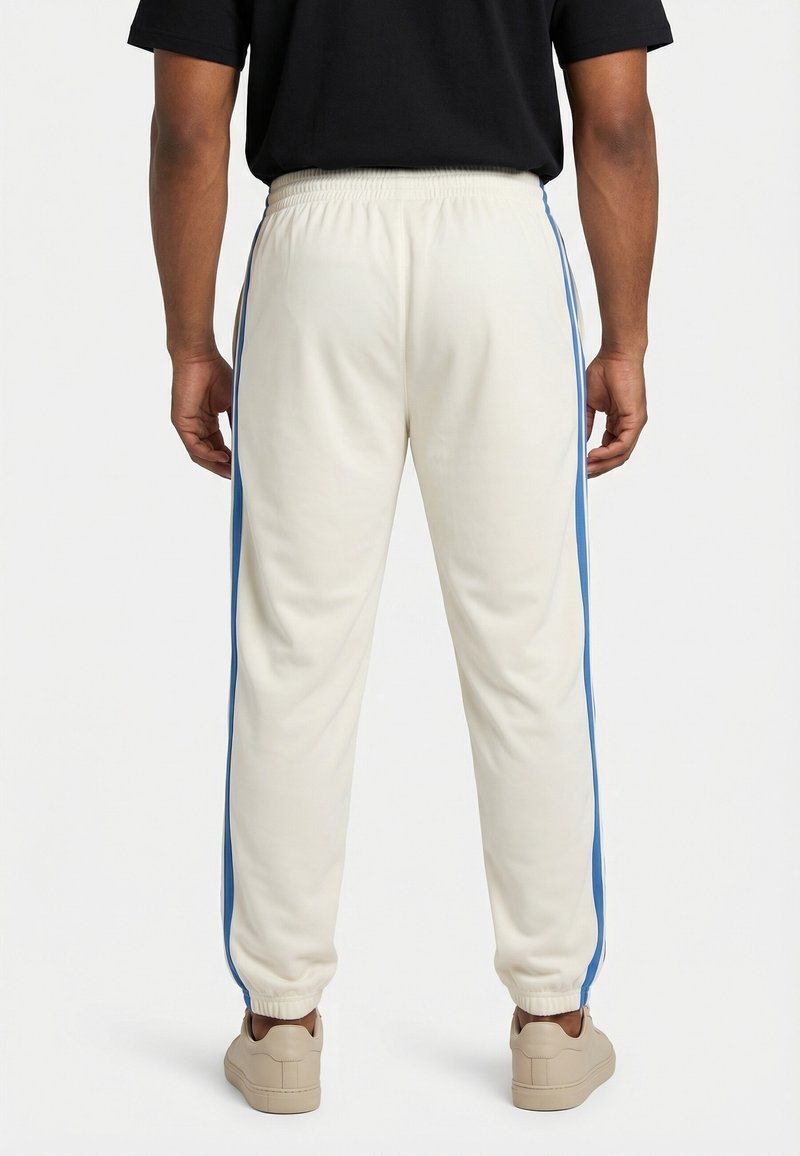 Uomo che indossa pantaloni jogger color crema con strisce laterali blu, scarpe da ginnastica beige e una maglietta nera, visto da dietro su sfondo bianco.
