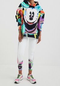 Färgglad hoodie med en grafik av en leende karaktär, i kombination med vita leggings som har olika livfulla mönster och accenter.