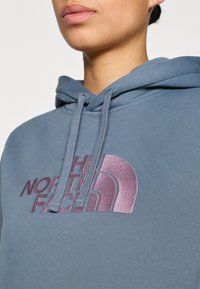 Φούτερ με κουκούλα από βαμβάκι σε ανοιχτό μπλε χρώμα με γκρι κορδόνια. Διαθέτει ένα εμφανές, λαμπερό μωβ λογότυπο τυπωμένο στο στήθος.