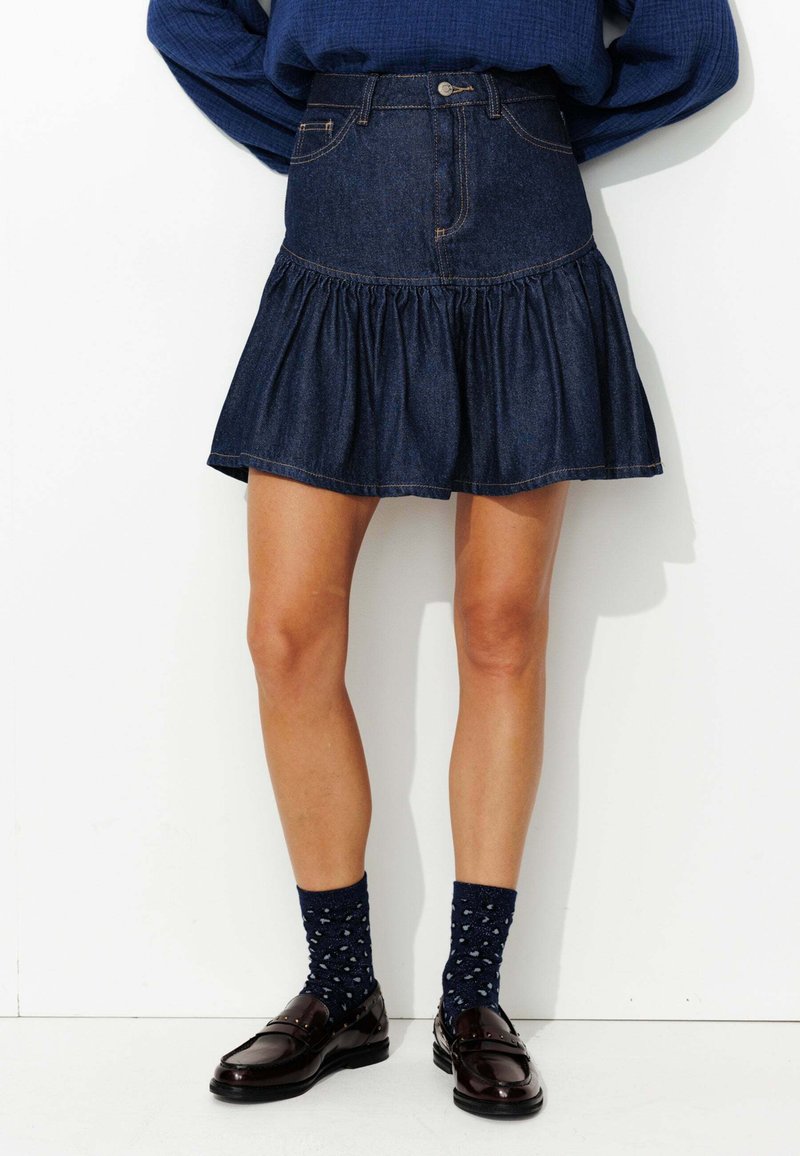Jupe en denim foncé avec un ourlet volanté évasé, dotée de poches avant et de passants de ceinture, associée à des chaussettes noires à motifs et des chaussures marron.