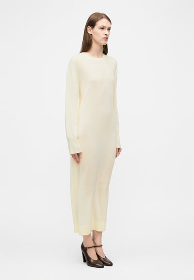 Γυναίκα που φοράει κρεμ μάλλινο φόρεμα με μακριά μανίκια και μήκος μέχρι τους αστραγάλους, συνδυασμένο με καφέ γόβες Mary Jane με ψηλό τακούνι, σε λευκό φόντο.