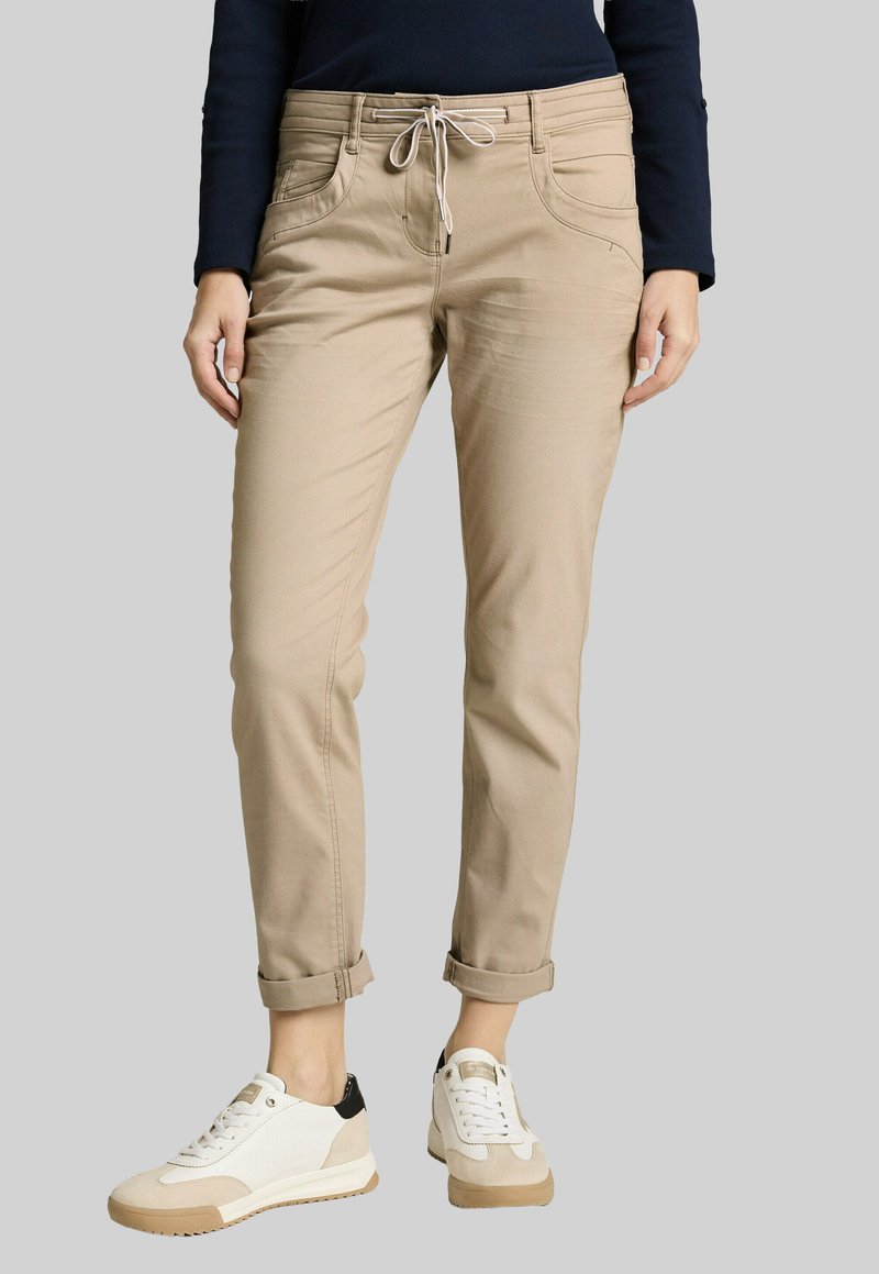 Beige cuffed pants with drawstring waist worn with white and beige sneakers and a dark long-sleeve top on a neutral background.