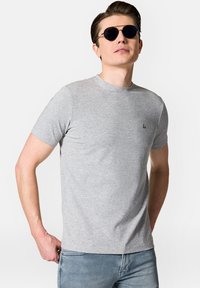 Szara koszulka z krótkim rękawem, uszyta z gładkiego materiału, z okrągłym dekoltem i małym czarnym logo na lewej piersi. Noszona z niebieskimi dżinsami.