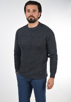 Suéter de punto azul marino texturizado con cuello redondo y puños acanalados, combinado con pantalones azules. Presenta un pequeño detalle del logo en el dobladillo.