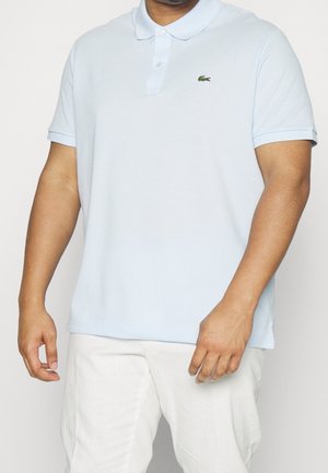 Homme portant un polo Lacoste bleu clair avec un pantalon blanc sur un fond uni.