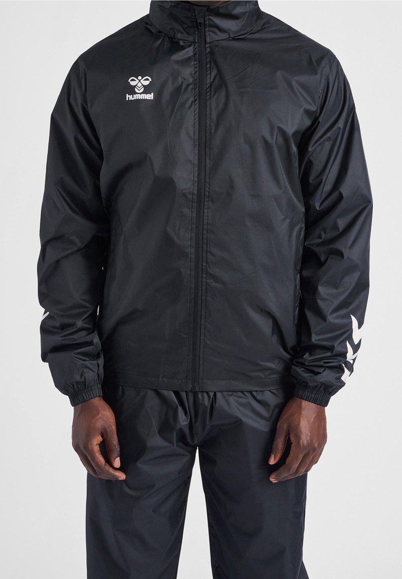 Veste de sport noire zippée intégralement avec capuche et logo blanc hummel sur la poitrine, motifs chevrons sur les manches, portée avec un pantalon noir assorti.