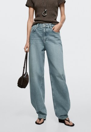 Personne portant un jean large bleu clair, un haut en tricot marron à manches courtes, des sandales compensées noires, portant un petit sac à main noir et un collier.