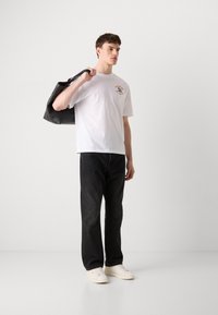 T-shirt bianco in cotone con grafica frontale, abbinato a jeans neri a vestibilità rilassata e sneakers bianche. Una borsa tote nera è appoggiata sulla spalla.