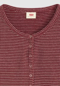 Top button-up a coste color bordeaux con scollatura rotonda e dettagli bianchi. Presenta cinque bottoni metallici lungo il davanti e un'etichetta al colletto.