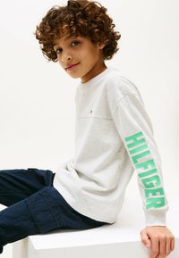 Lys grå langærmet skjorte med en grøn "HILFIGER" print på venstre arm, båret med mørkeblå bukser og med et lille logo på brystet.