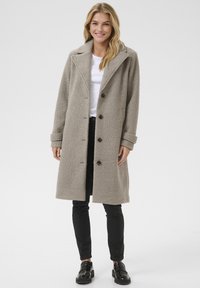 Lichtgrijze, textuurrijke jas met een reverskraag, sluiting met knopen aan de voorkant en zijzakken, gecombineerd met zwarte broek en glanzende zwarte schoenen.