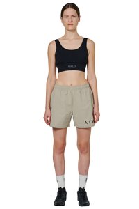 Kobieta w beżowych szortach z elastycznym pasem z napisem "ATW", czarnym sportowym staniku z logo "HALO", białych skarpetkach i czarnych butach sportowych, stojąca prosto.