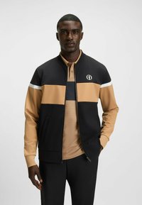 Black and tan bomber jacket with a grid texture, white accents, and a logo on the chest. Worn over a light brown collared shirt.