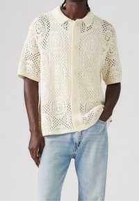 Man in een crème korte mouw haakgebreide cardigan met kraag en knopen, gecombineerd met lichtblauwe jeans, hand in de zak.