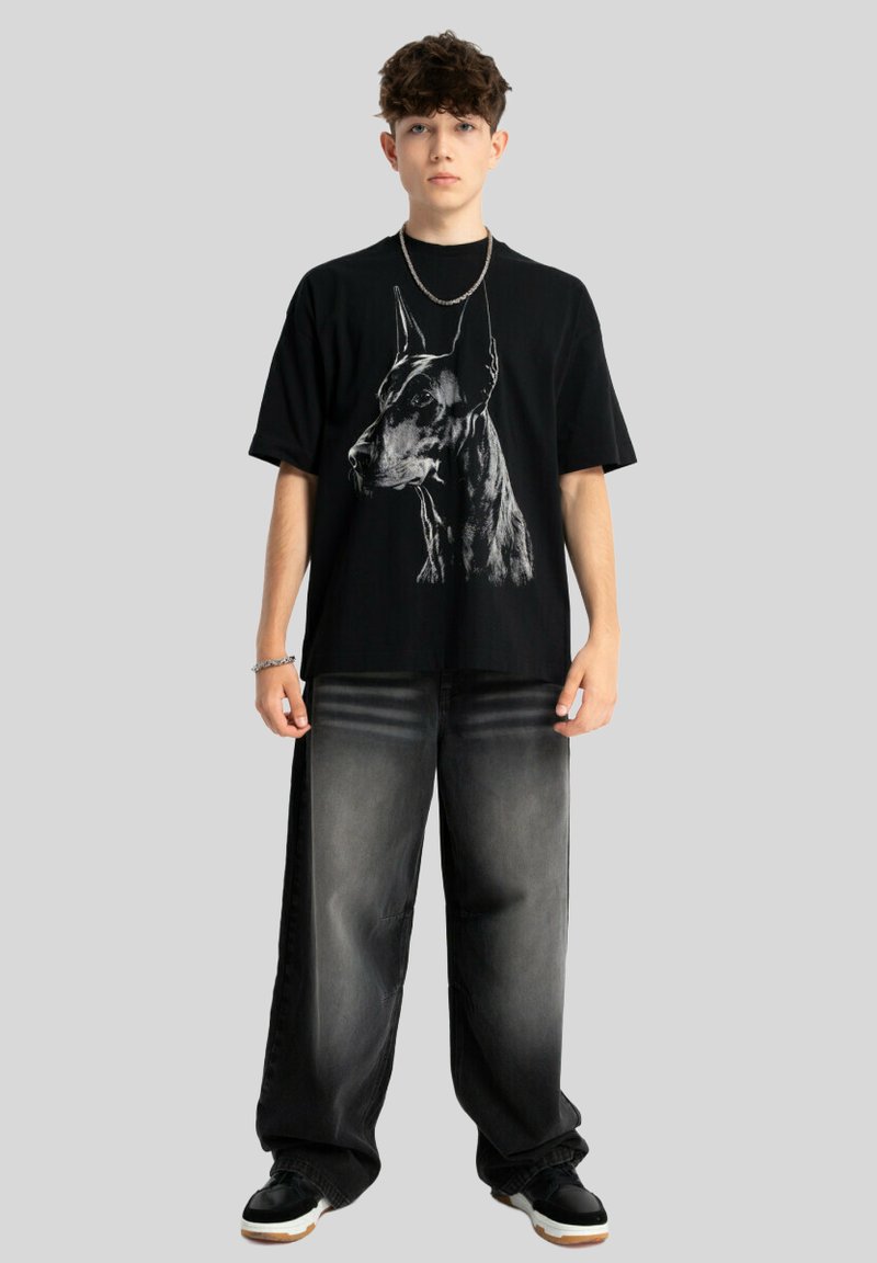 Czarny graficzny t-shirt z motywem głowy psa, zestawiony z luźnymi, szarymi dżinsami i czarnymi trampkami z białymi akcentami.